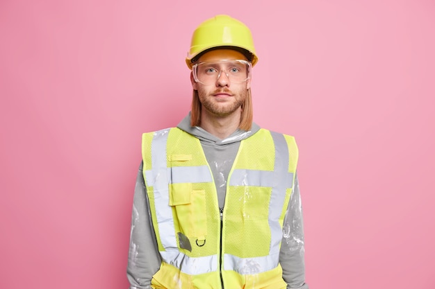 Free photo serious man builder engineer wears construction safety helmet uniform glasses looks confidently  ready for work isolated over pink wall. self assured workman or construction worker