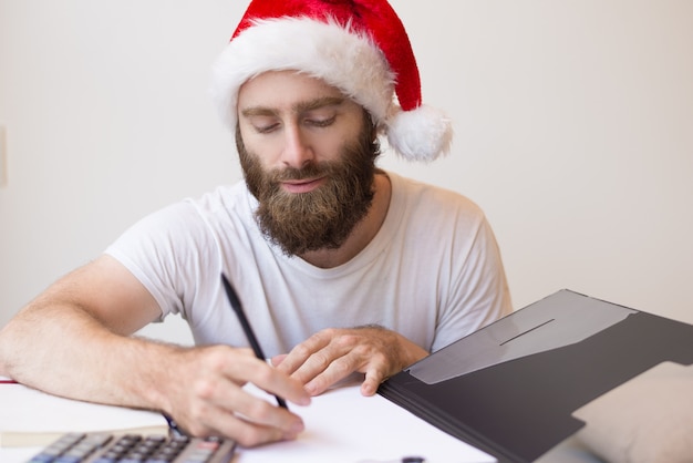 Serious man wearing Santa hat and working with documents