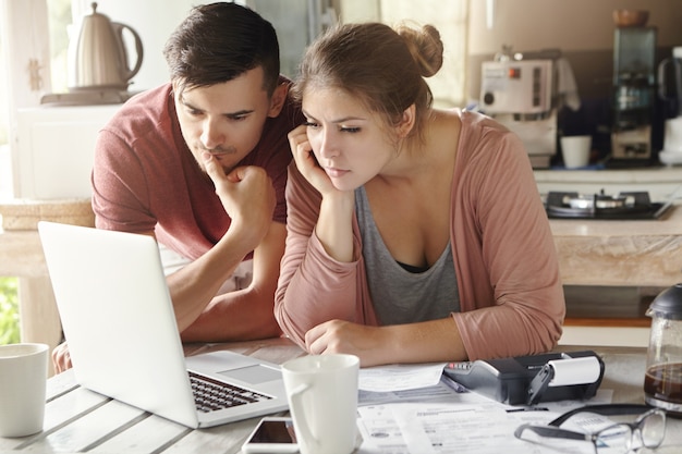 Serious man and woman sitting at kitchen table in front of open laptop