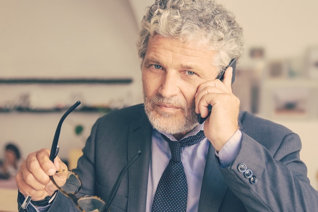 Free Photo serious mature businessman talking on mobile phone, standing at co-working, leaning on desk