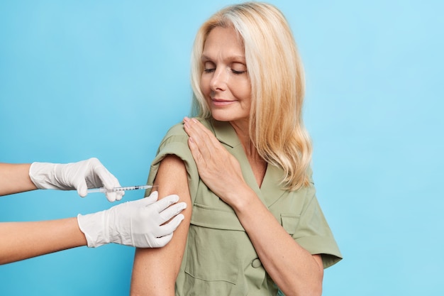 Free Photo serious middle aged woman with fair hair looks attentively at process of vaccination gets shot in shoulder to protect herself from coronavirus isolated on blue