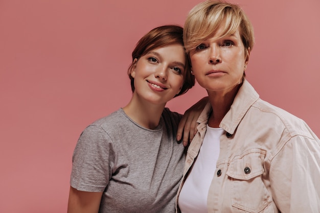 Free photo serious old woman with blonde hair in cool jacket looking into camera together with smiling girl with brunette hair in grey t-shirt on pink backdrop.