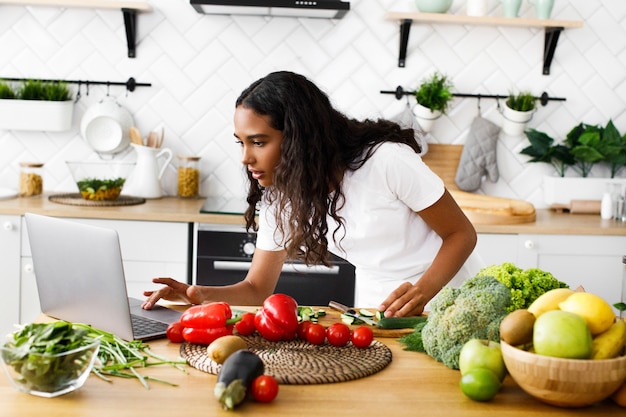 Serious pretty mulatto woman is looking on the laptop screen  on the modern kitchen on the table full of vegetables and fruits, dressed in white t-shirt