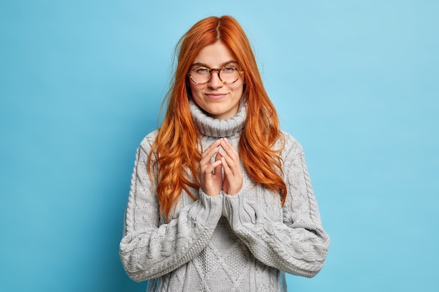 Serious redhead young European woman schemes something steepls fingers and looks with intention to do wears optical glasses grey knitted sweater.