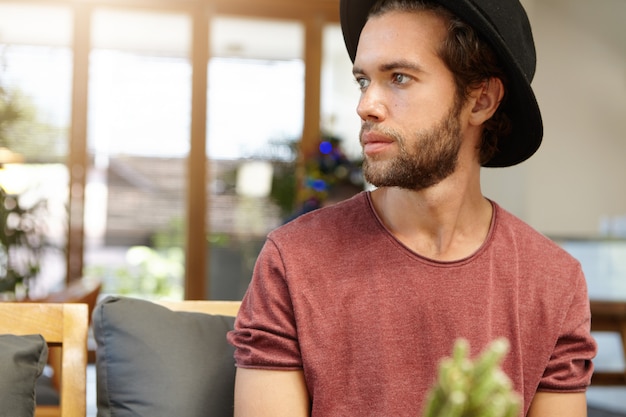 Free photo serious or sad attractive young bearded student wearing trendy black hat sitting alone at modern spacious coffee shop