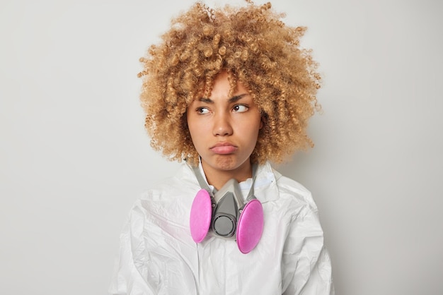 Serious sad curly woman wears chemical protective suit and gas mask looks away with dissatisfied expression frustrated because of biohazard isolated over grey background Radiation protection