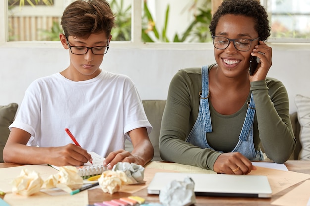 Serious schoolboy in white t shirt, writes down records in notepad, busy with studying