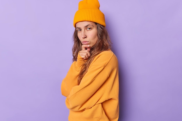 Free Photo serious self confident beautiful teenage girl holds chin looks directly at camera considers something stands sideways against purple wears stylish hat and jumper. human face expressions