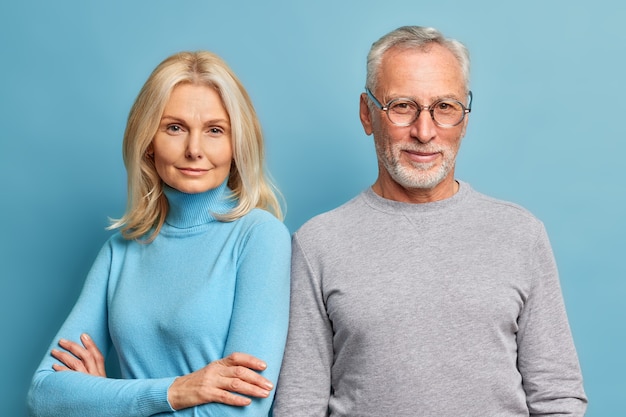 Serious self confident blonde pretty woman stands with arms folded near her husband pose for making common photo dressed in casual turtlenecks isolated over blue wall