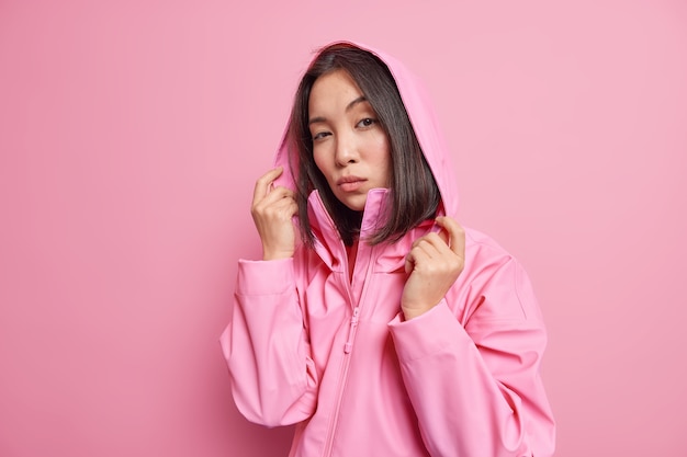 Free photo serious self confident millennial girl with dark hair wears anorak hood on head looks directly  models against pink wall going to have walk during cold windy day. people and style