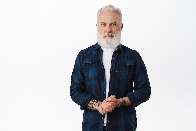 Serious senior man hold hands together standing in content pose ready to help how may I assist you posing selfassured and determined against white background