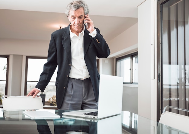 Free Photo serious senior man talking on mobile phone with laptop and digital tablet on reflective desk