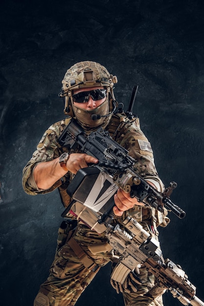 Serious soldier in full gear and military uniform is standing with machine gun over dark background.