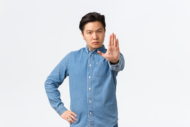 Serious strict asian man extending hand to shop stop gesture, scolding person or disagree, prohibit action, forbid doing something bad, standing over white background and give warning.