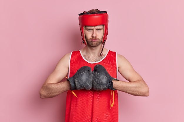 Free Photo serious strong unshaven boxer wears hat and boxing gloves keeps hands together prepares for sport competition ready to fight.