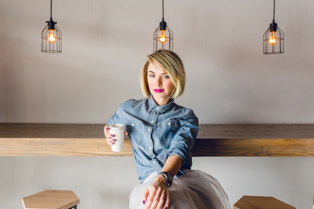 Serious stylish girl with blonde hair and pink lips sitting in a coffee shop with wooden chairs and table. She holds a cup of cofee