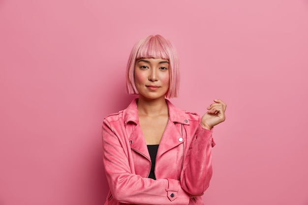 Free photo serious stylish lady looks with calm expression direcrly, has healthy skin, rouge cheeks, wears pink hair wig, dressed in jacket, keeps hand raised,