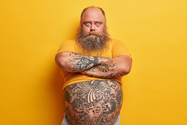 Serious surprised blue eyed man with thick beard, keeps arms folded, listens wife explanation, feels jealous, has big tummy, overweight because of wrong nutrition, isolated on yellow wall