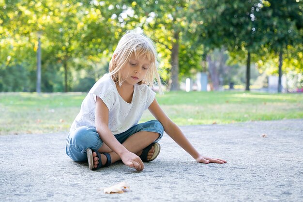 Serious sweet fair haired girl sitting and drawing with colorful pieces of chalks. Copy space. Childhood and creativity concept