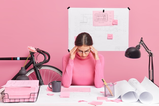 Free photo serious talented young european female architect being busy at her workplace makes project of new residential complex concentrated in papers wears casual turtleneck tries to gather with thoughts