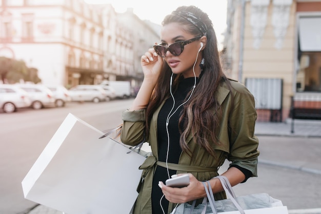 Free Photo serious tanned woman in dark glasses listening music in white earphones. charming hispanic lady in trendy jacket carrying purchases, walking on the street.