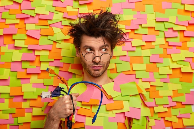 Free photo serious thoughtful man holds bunch of colorful cables, going to repair computer, has messy hairstyle, looks through optical glasses, poses against wall with stickers. technology, engineering