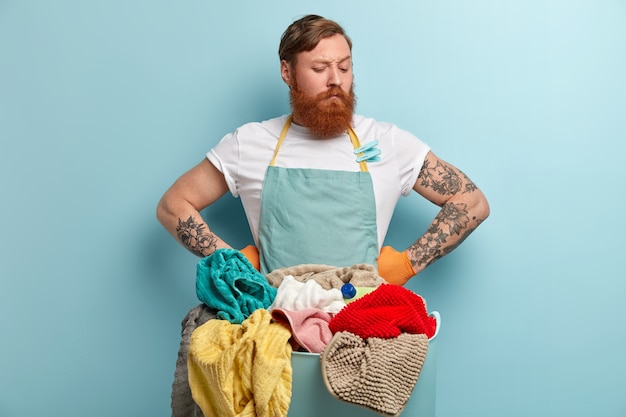 Free photo serious thoughtful redhead man holds both hands on waist, has thick bristle, looks down, wears casual t shirt and apron, stands in front of basin with laundry and cleaning supply isolated on blue wall