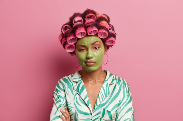 Free photo serious unimpressed woman stands with crossed hands over body, feels bored during beauty procedures, wears hair rollers for creating perfect curls, green mask to rejuvenate and reduce wrinkles