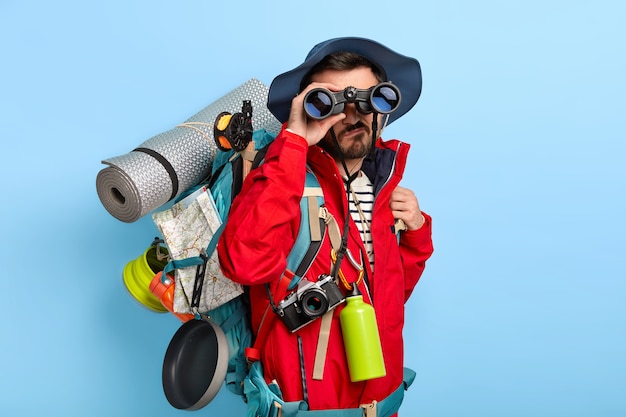 Free Photo serious unshaven male backpacker keeps binoculars near eyes, wears hat and red jacket, explores new way, carries tourist backpack