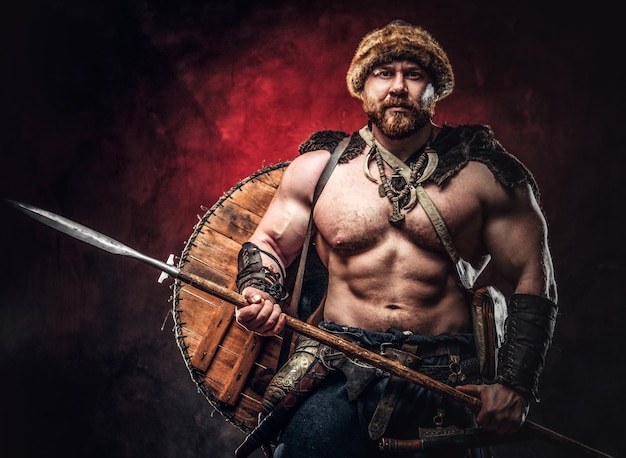 Free Photo serious viking clad in light armor with a shield behind his back holds a spear. posing on a dark background with red light