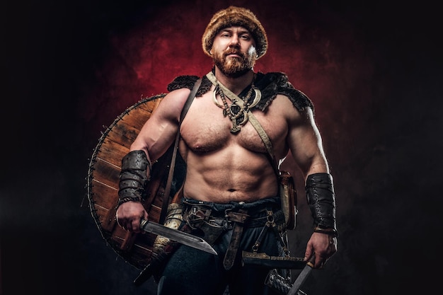 Serious Viking clad in light armor with a shield behind his back holds a sword and an axe. Posing on a dark background with red light