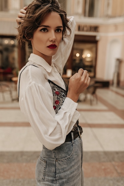 Free Photo serious woman in white shirt and jeans with black belt looking away in city. brunette woman with bright lips posing at street.