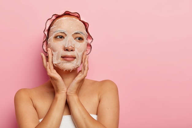 Serious woman with bare shoulders applies moisturising paper mask on face, touches cheeks gently, has soft healthy skin, wears bathcap