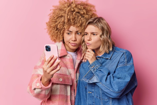 Free photo serious women look attentively at smartphone screen make photo or make video call wear fashhionable clothes stand closely to each other isolated over pink background people technology concept