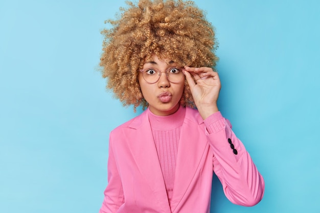 Free photo serious wondered woman with curly bushy hair keeps lips folded hand on rim of spectacles dressed in formal outfit reacts on shocking news isolated over blue background human face expressions