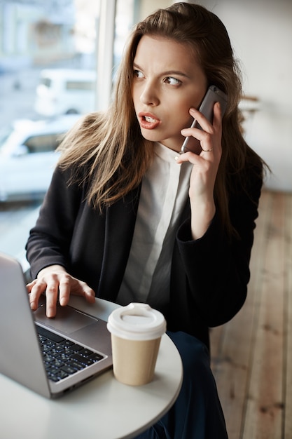 serious worried european woman sitting in cafe, drinking coffee and working with laptop, talking on smartphone while looking aside anxiously