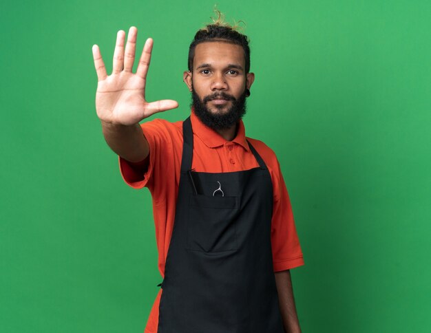 serious young afro-american male barber wearing uniform doing stop gesture isolated on green wall with copy space