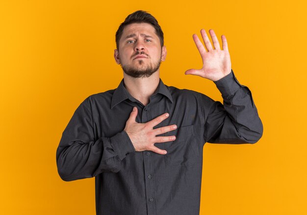 Serious young blonde handsome man keeping hand on chest and up other empty hand looking at camera 