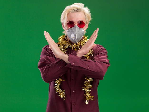 Free Photo serious young blonde man wearing glasses and protective mask with tinsel garland around neck  doing no gesture isolated on green wall