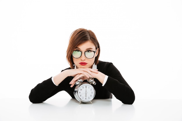 Free photo serious young business lady wearing glasses with alarm clock