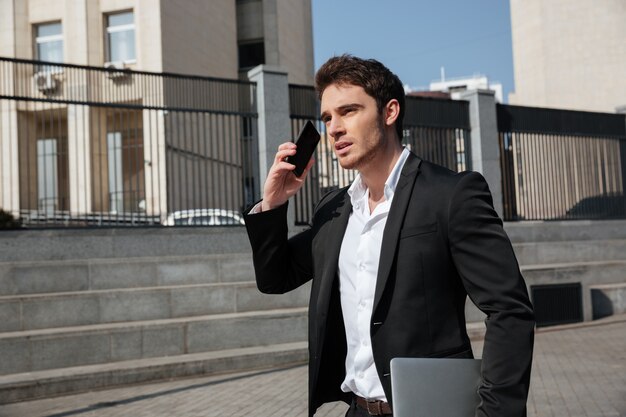 Serious young businessman walking outdoors talking by phone.