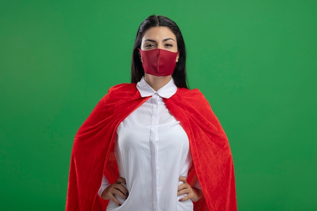 Free photo serious young caucasian superhero girl wearing mask holding hands on her hips looking at camera with serious look isolated on green background with copy space