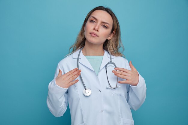 Serious young female doctor wearing medical robe and stethoscope around neck pointing at herself with hands 