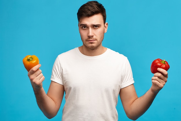 Serious young guy with bristle holding orange and red bell peppers with indecisive facial expression, having doubts and suspicions, does not want to eat pesticidal vegetables