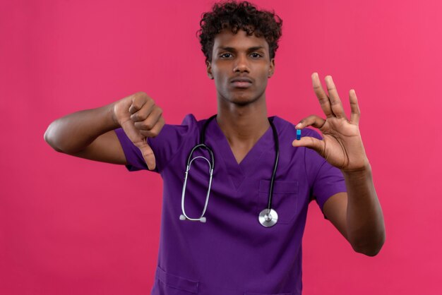 A serious young handsome dark-skinned doctor with curly hair wearing violet uniform with stethoscope showing thumbs down while holding pill 