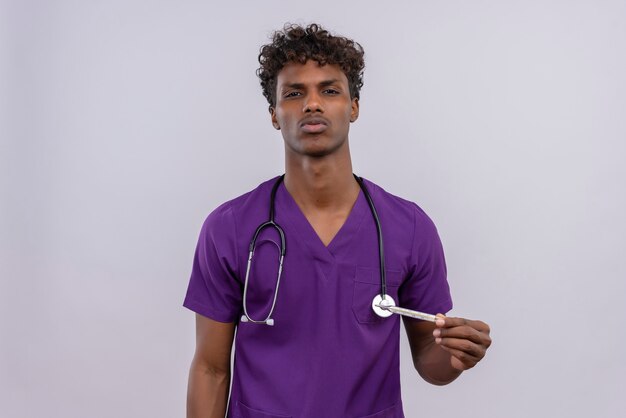 A serious young handsome dark-skinned doctor with curly hair wearing violet uniform with stethoscope  while holding thermometer 