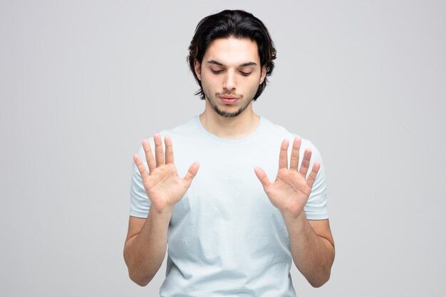 Serious young handsome man looking down showing stop gesture with both hands isolated on white background