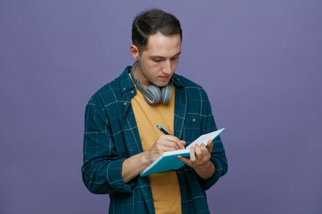 Serious young male student wearing headphones around neck writing with pen on note book isolated on purple background