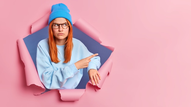 Serious young redhead woman pointing at copy space, wears blue sweatshirt and hat. breaks through pink paper wall.