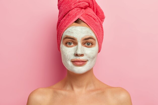 Serious young woman with clay facial mask, wears wrapped towel, nourishes skin with vitamins